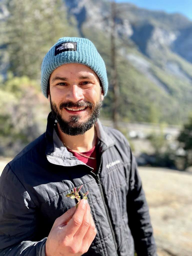 Smilling man with light-tan complexion and dark beard, holding a Mimulus flower the mountainous outdoors